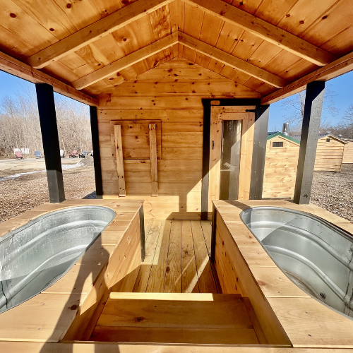 A wooden outdoor structure with two built-in bathtubs, one on each side, and a small enclosed room at the back under a peaked wooden roof.