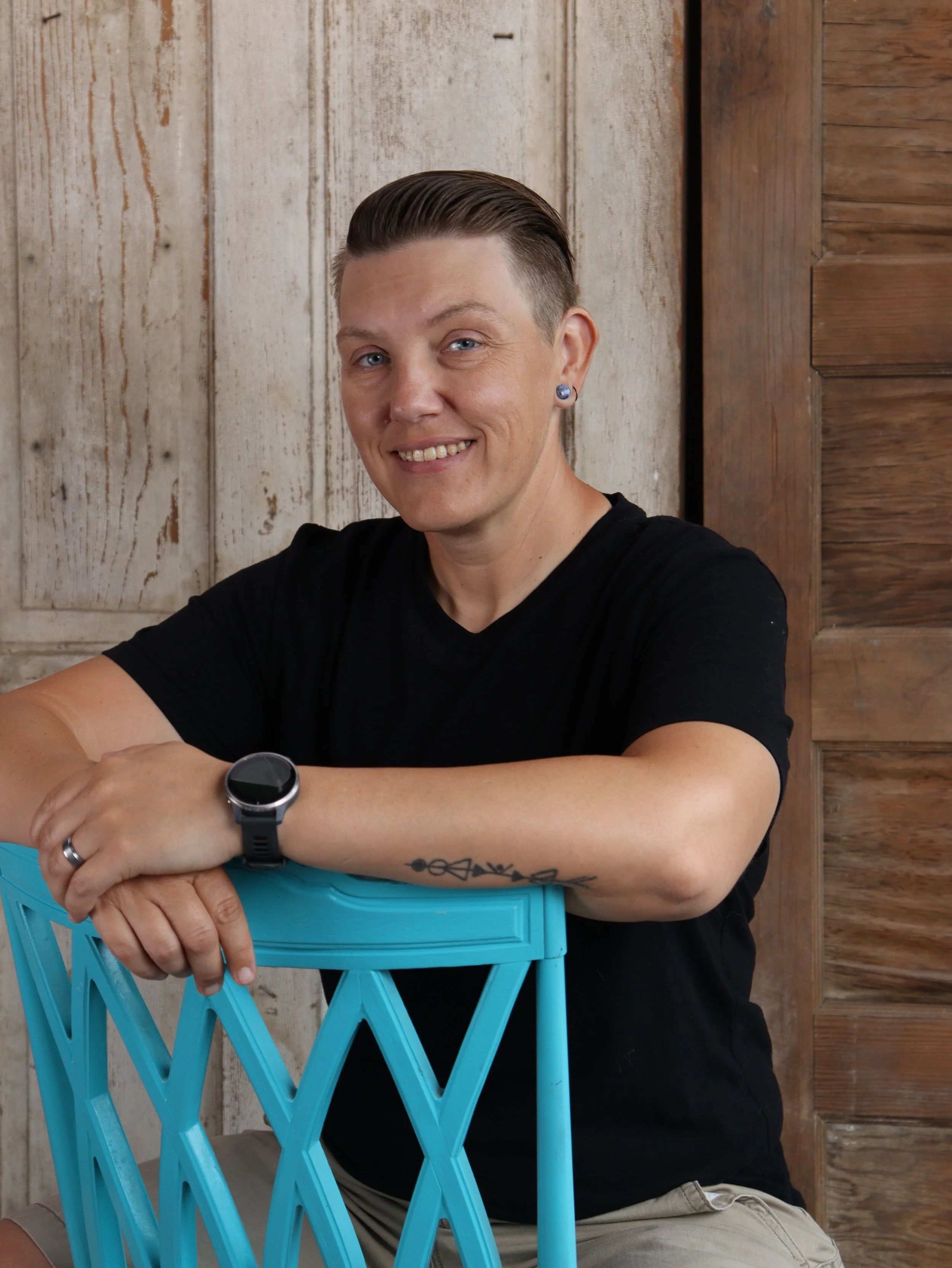 A woman with short slicked-back hair, wearing a black shirt, beige shorts, and a wristwatch, smiling and leaning on a blue plastic chair, seated against a wooden wall background.