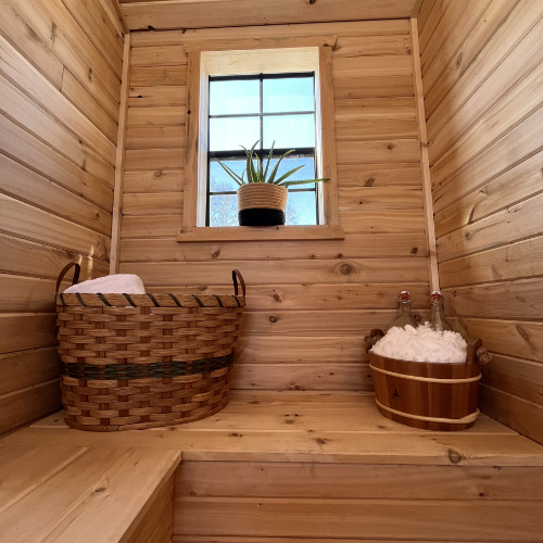Small wooden room with a window, a potted plant, a woven basket with a towel, and a wooden tub filled with bubbles.