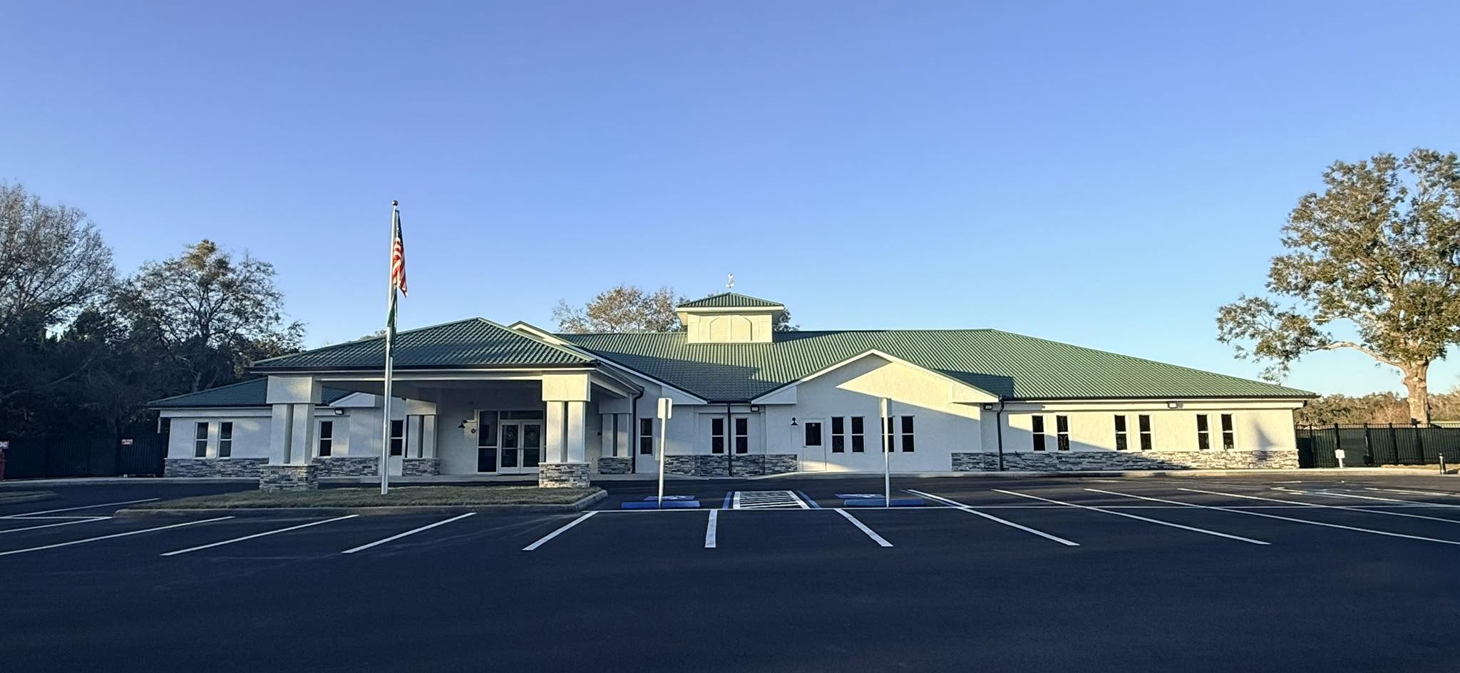 A single-story building with a green metal roof, white walls, and stone accents at the base. There is an American flag on a flagpole in front, an empty parking lot, and a few trees around the building.