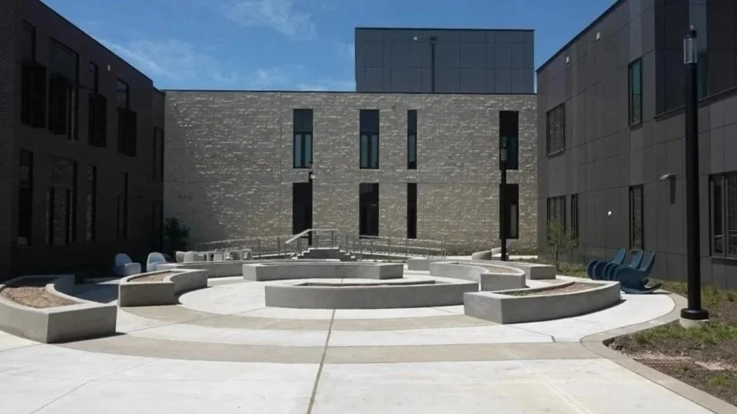 A modern outdoor courtyard with concrete seating and benches, black lampposts, and blue chairs, surrounded by contemporary buildings.