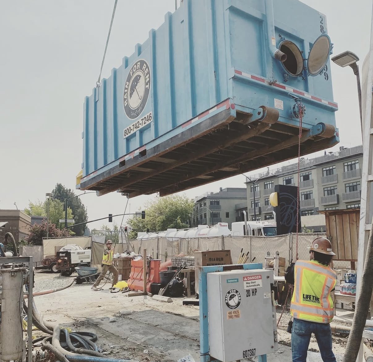 Construction site where a large blue shipping container is being hoisted overhead by lifting equipment. Several workers are present, wearing safety gear, and there are various construction materials and equipment around.
