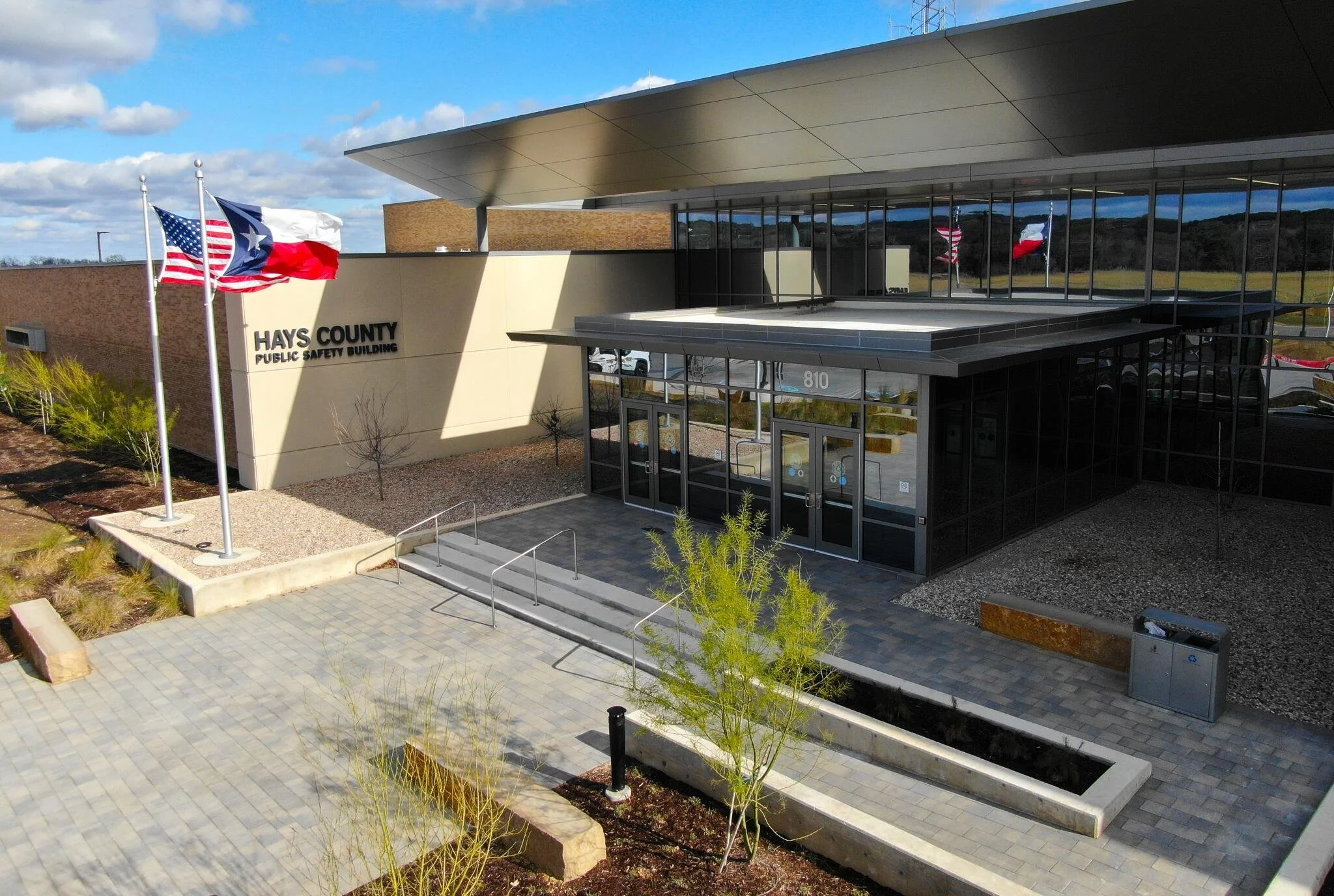 Exterior view of Hays County Public Safety Building with flags in front, glass entrance, and a parking lot.