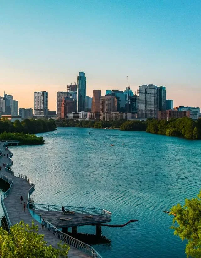 View of a city skyline with tall buildings across a wide river, with a promenade and green trees in the foreground during sunset.