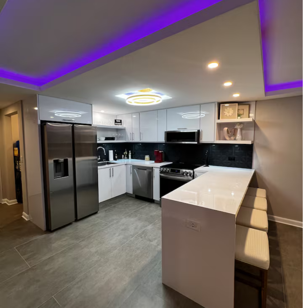 Modern kitchen with white cabinets, stainless steel refrigerator, black backsplash, and bar seating with three beige stools. Purple and yellow lighting accents the ceiling.