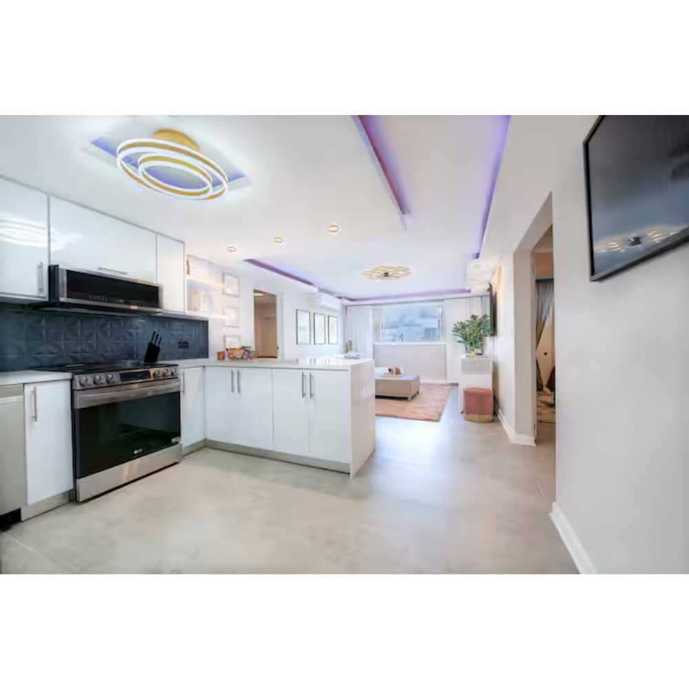 Modern white kitchen with black backsplash, stainless steel appliances, and purple LED ceiling lights, leading into a bright living room with a beige sofa and large window.