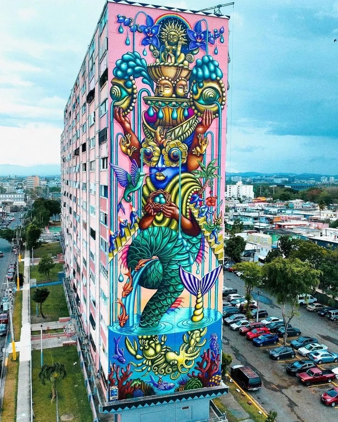 Colorful mural covering the side of a tall building, depicting a fantastical creature with a human face, a mermaid tail, and elements of nature such as trees, flowers, and a hummingbird.