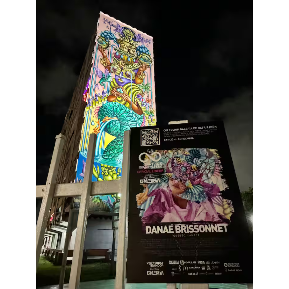 A tall, illuminated mural with colorful, abstract, and fantastical designs on a building, and an informational poster featuring Danaé Brissonnet in front of it, at night.