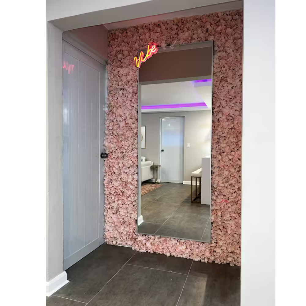 A large mirror with a pink flower wall frame, a small neon sign that says 'Vibe,' and a view into a room with white walls, gray tiled floors, a white door, a white chair, and purple LED lighting.