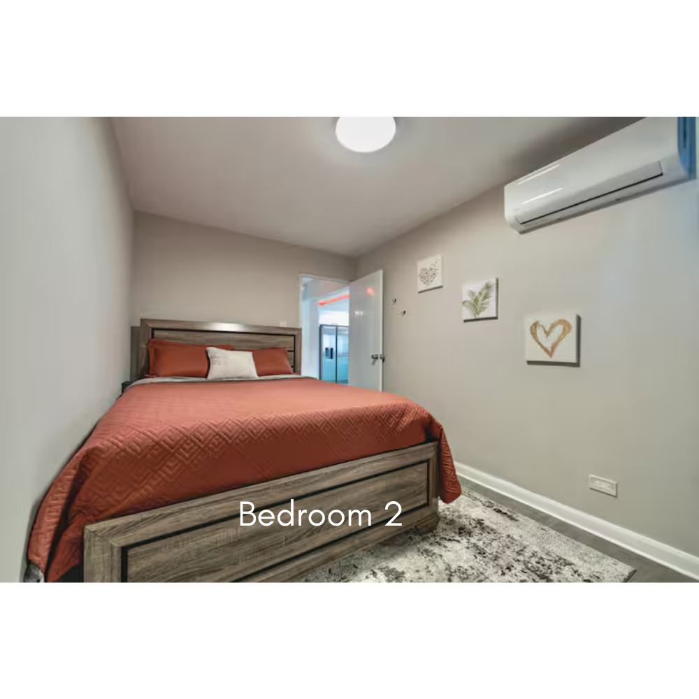 A cozy bedroom with a wooden bed frame, rust-colored quilt, and white pillows in Bedroom 2. The wall has three decorative art pieces, an air conditioning unit, and an open door leading to another room or balcony.