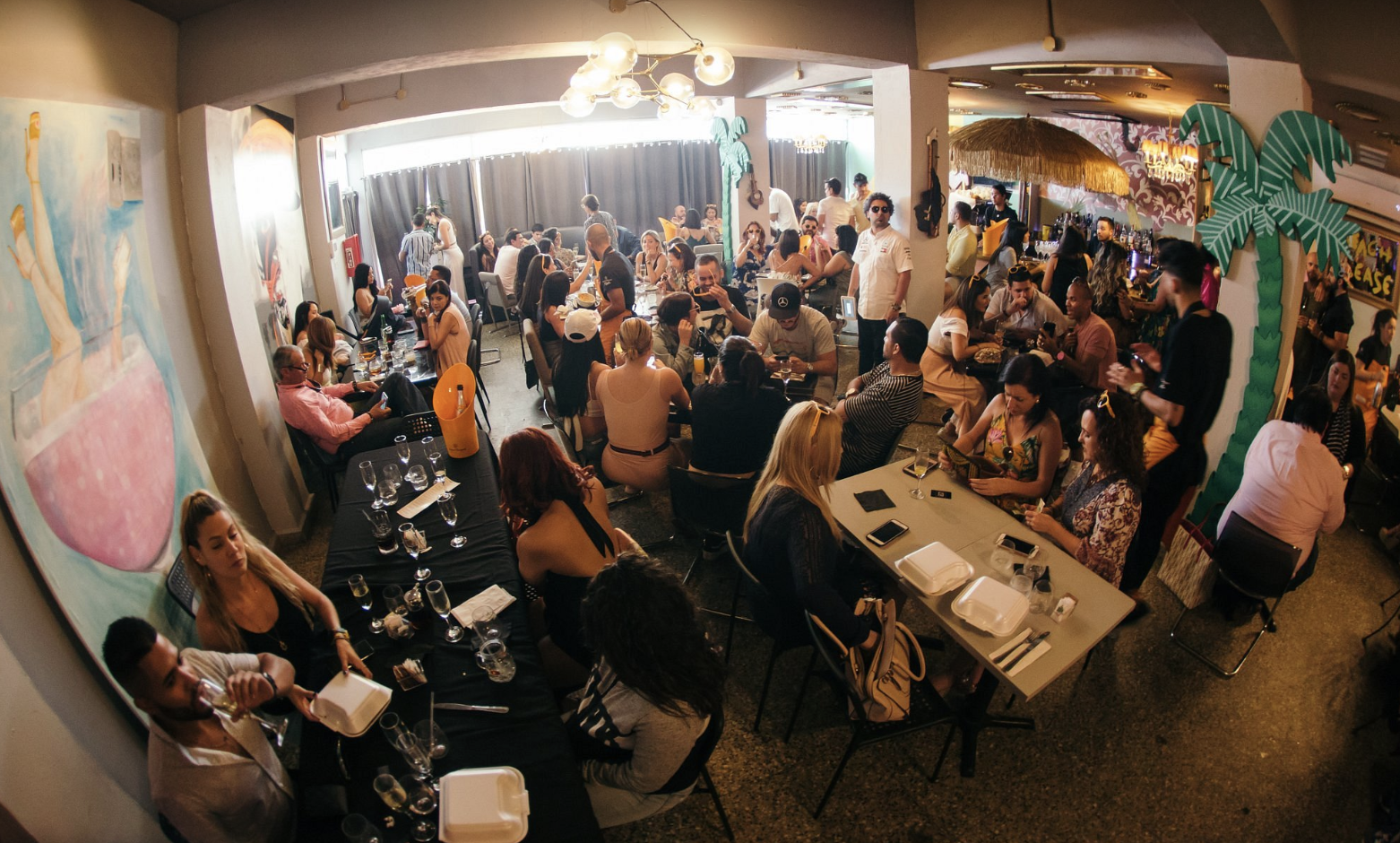Crowded restaurant with people dining and socializing, tropical-themed decor, paintings, and bright lighting.
