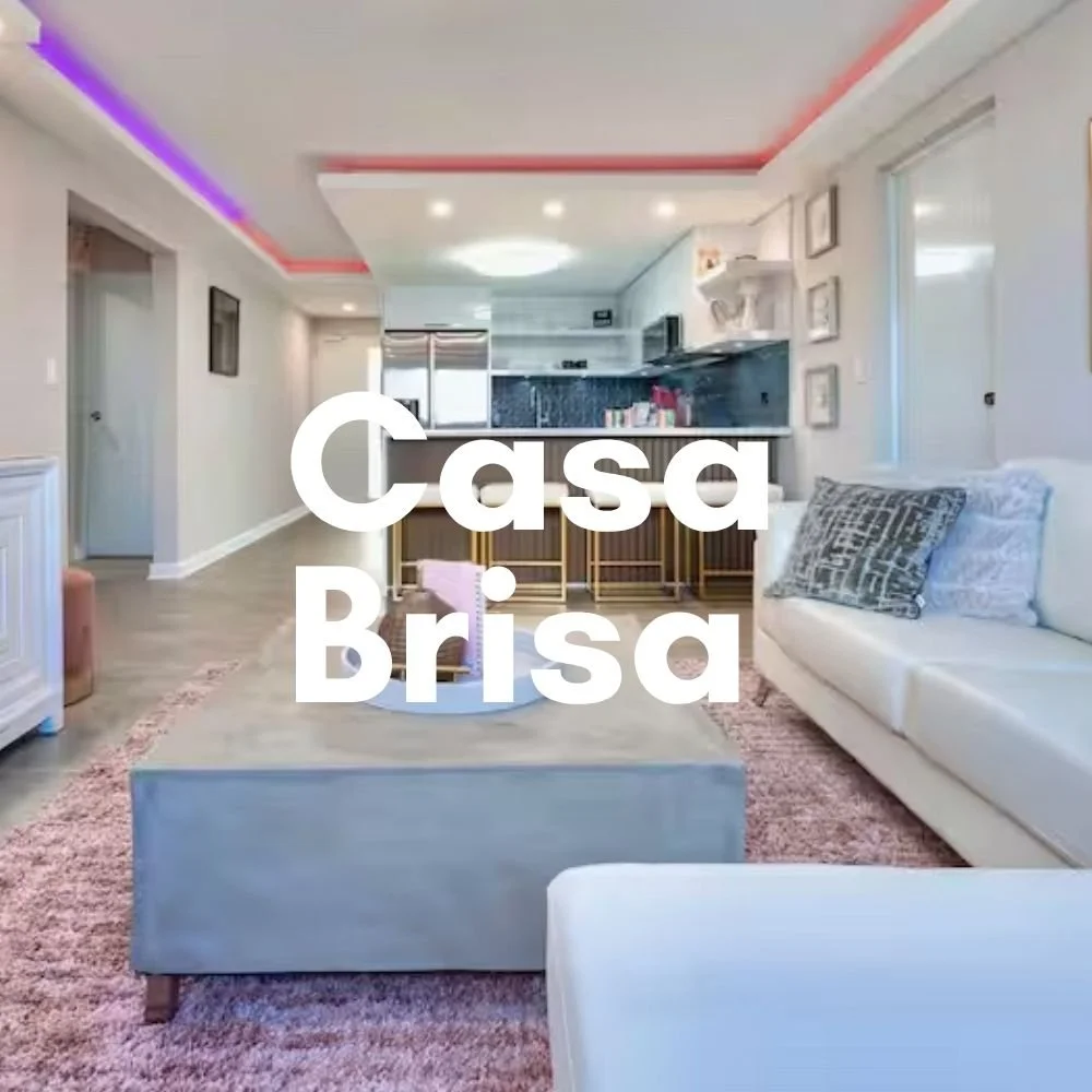 Living room with white sofa, pink carpet, and open kitchen area in the background, featuring a black backsplash and kitchen island with bar stools. Text overlay says 'Casa Brisa'.