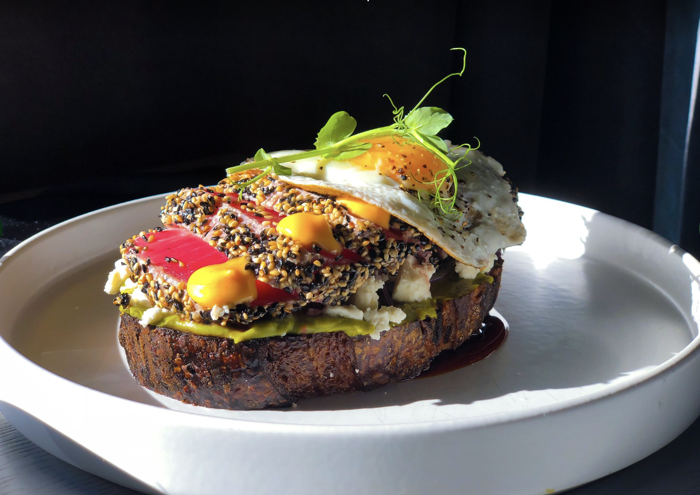 A toasted bread topped with avocado, sliced tomato, a fried egg, microgreens, and sesame seeds, drizzled with yellow sauce, on a white plate.