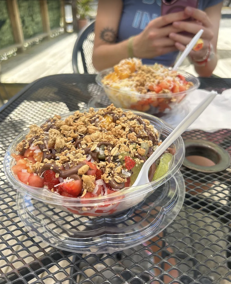 Two bowls of fruit salad topped with granola and chocolate drizzles sit on a black metal outdoor table. A person in a blue shirt is taking a photo of the bowls with a pink phone in the background.