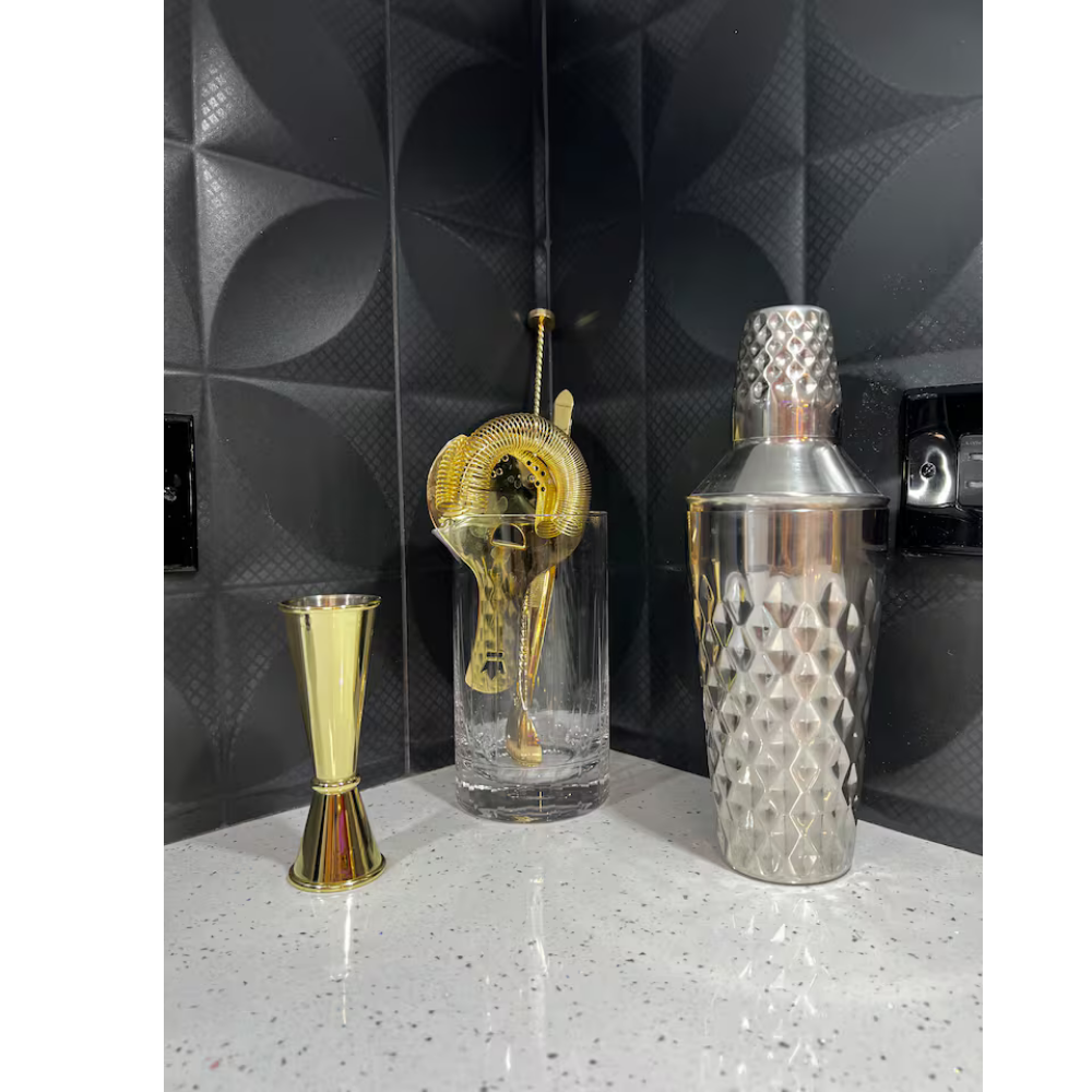 Gold cocktail jigger, crystal glass with gold cocktail strainer, metallic cocktail shaker on speckled white countertop with black textured wall background.