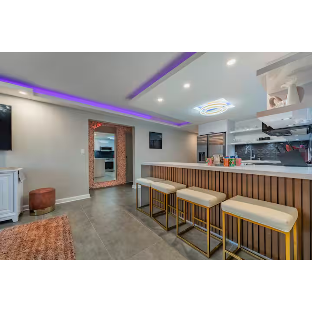 Modern kitchen and bar area with four cream-colored bar stools with gold frames, a white countertop, microwave, and decorative items on the counter, recessed lighting, and a purple LED light strip on the ceiling.