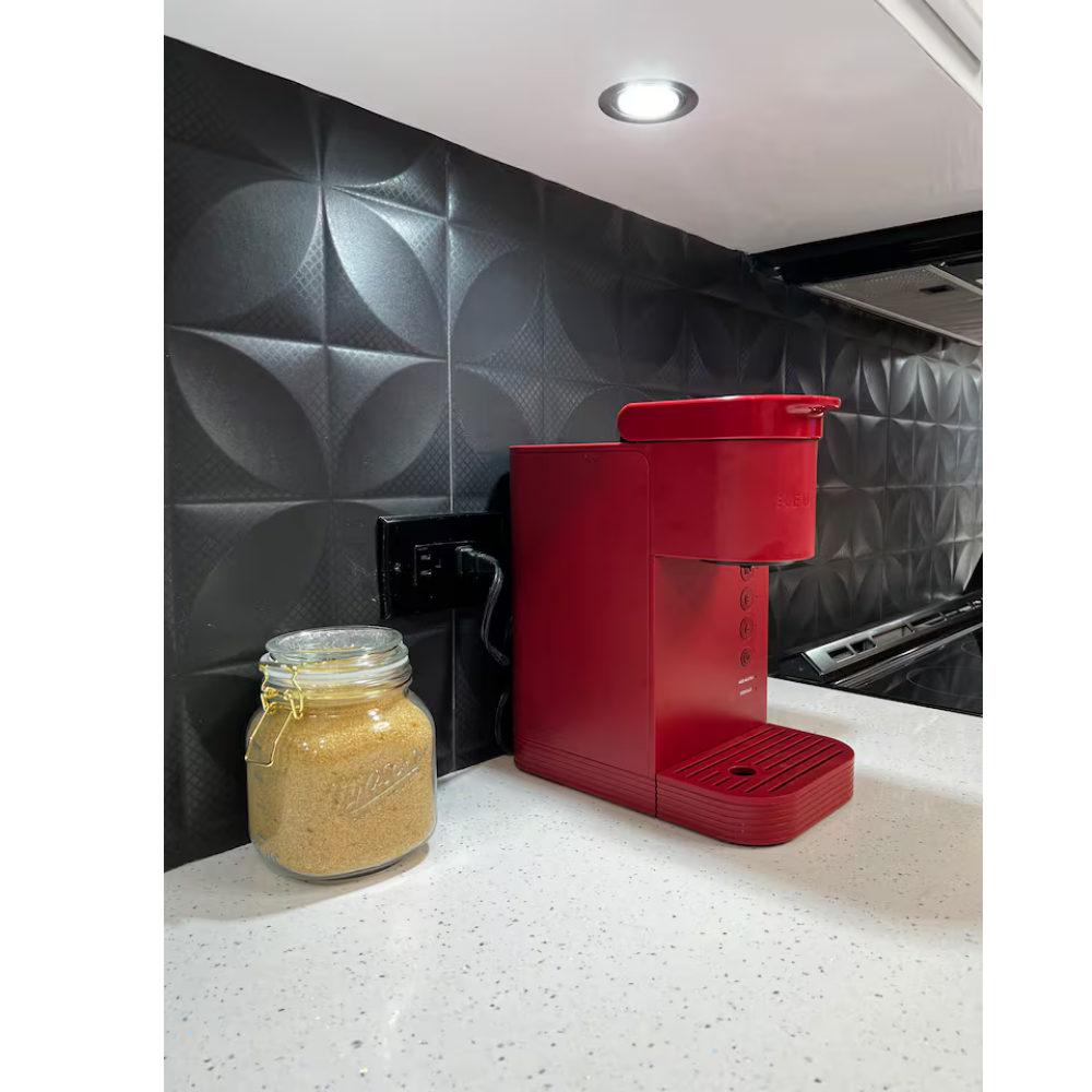 A red coffee maker and a jar of brown sugar on a white countertop in front of black textured tile wall with circular patterns.