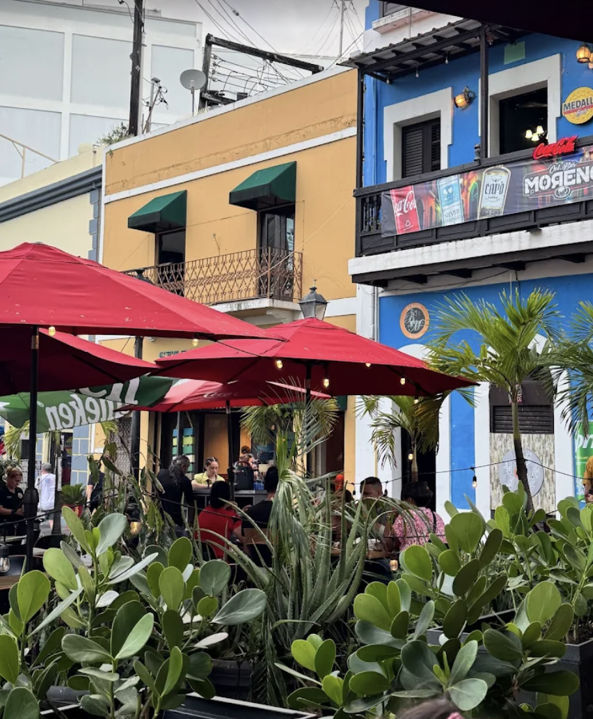 Outdoor restaurant patio with red umbrellas, surrounded by green plants, with people dining. Buildings painted in bright colors, including yellow, blue, and black in the background.