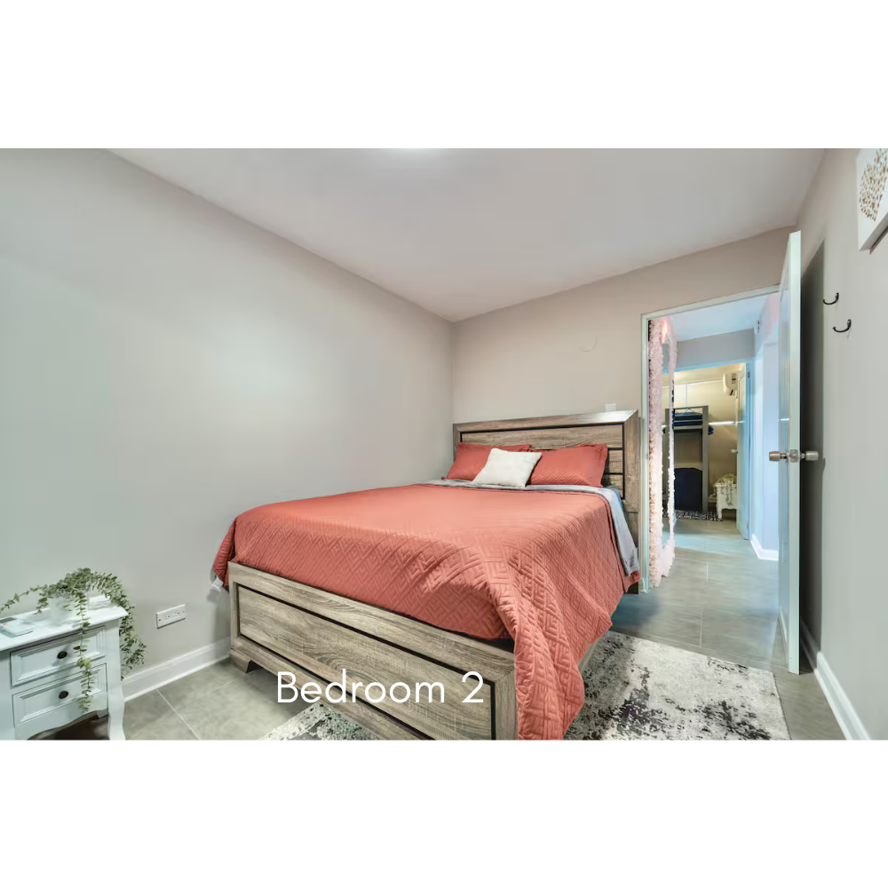 Bedroom with a bed covered in a coral-colored quilt and a white pillow, small white nightstand with greenery, gray walls, wooden floor, doorway leading to another room.