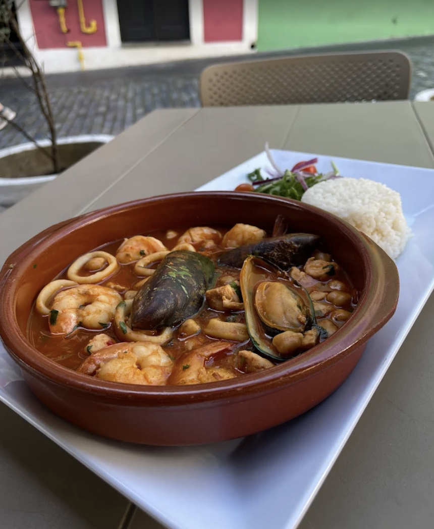 Seafood stew with shrimp, mussels, and squid, served in a clay dish, with a side of white rice and salad on a white plate.