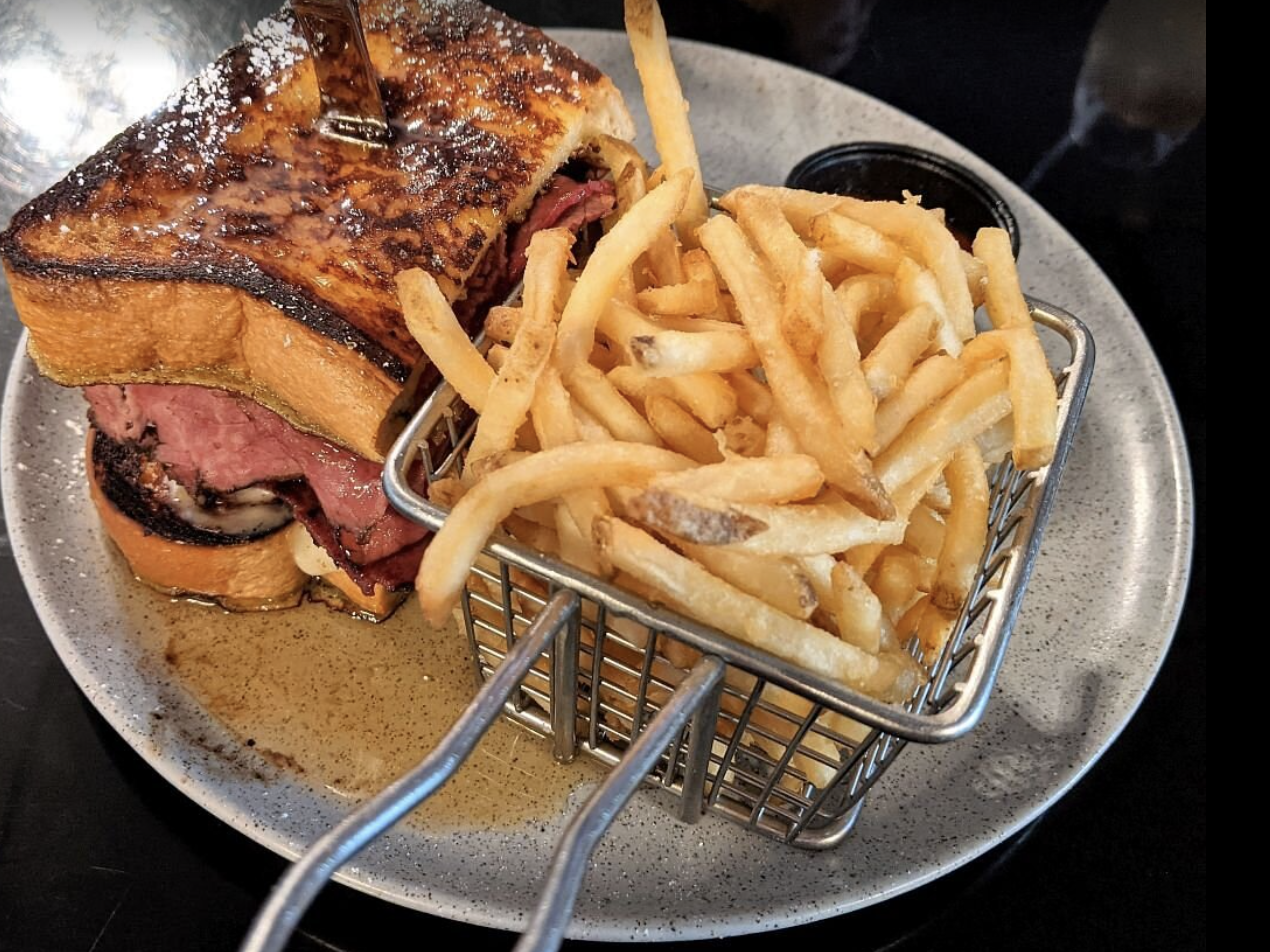 A plate with a grilled steak sandwich with melted cheese, served with a side of French fries, and a small container of dipping sauce.