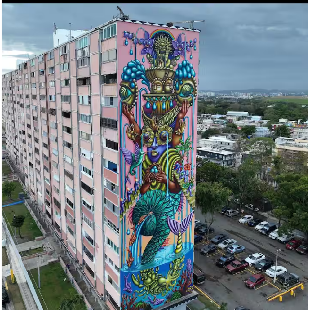 Colorful mural on side of a pink apartment building depicting a mystical sea creature with a mermaid tail, surrounded by vibrant marine life, flowers, and abstract elements.