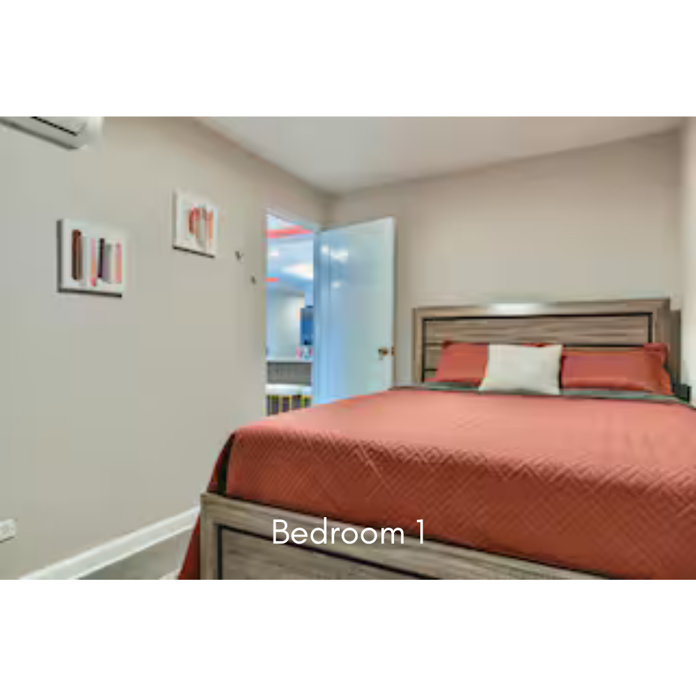 A bedroom with a bed covered in a rust-colored quilt, wooden headboard, and a white pillow. The wall has two small abstract framed artworks, an open door leading to another room, and a small window with a view of the outside.
