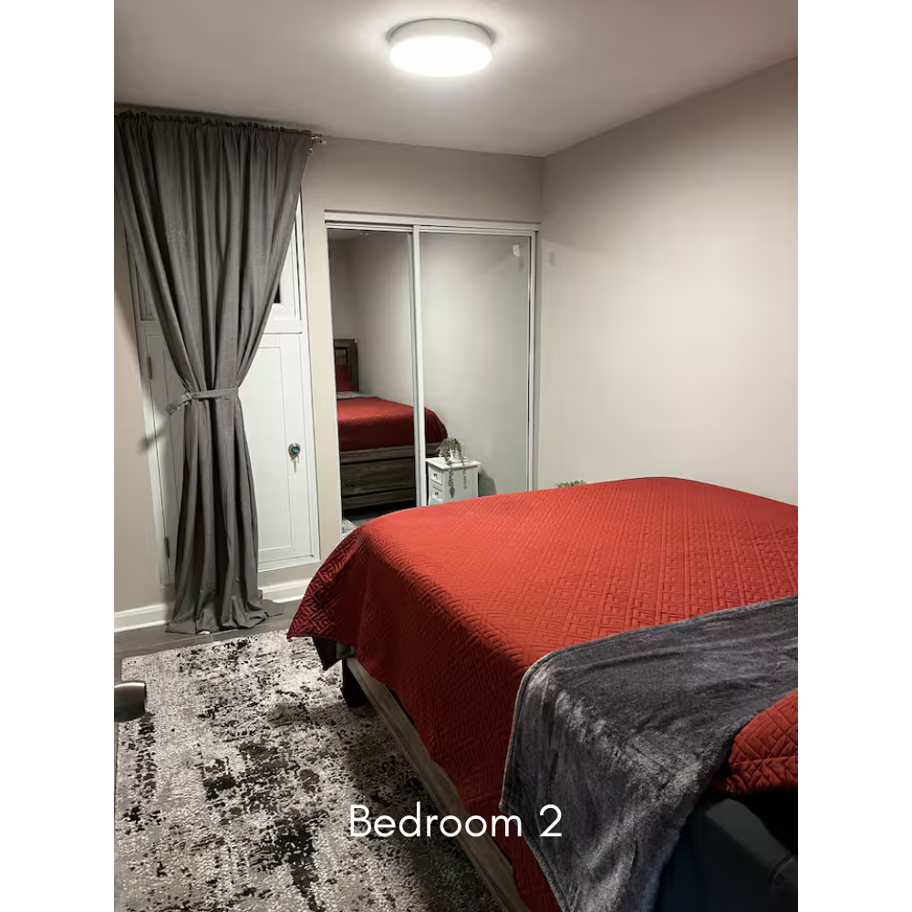 A bedroom with a bed covered in a red bedspread, a mirrored closet, a nightstand with decorative items, a dark gray blanket on the bed, and a large gray curtain next to a window. Label reads "Bedroom 2."