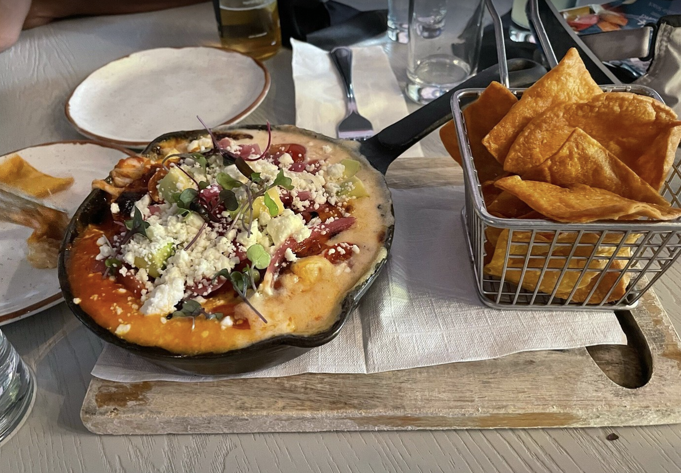 A pizza topped with cheese, vegetables, and microgreens on a wooden serving board with a side of tortilla chips in a basket.