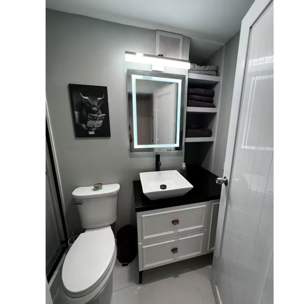 Small modern bathroom with a white toilet, black vanity with a white vessel sink, illuminated mirror, and open shelves with dark towels.