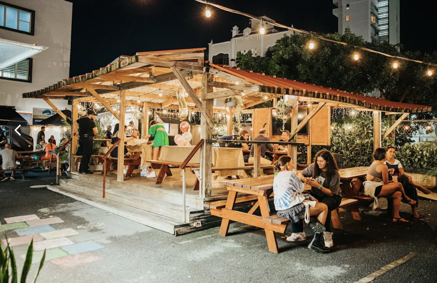 Nighttime outdoor patio dining area with people sitting at tables, string lights overhead, and a wooden structure with a red roof.