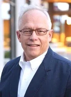 Tim Reeves, pictured with glasses short gray hair, wearing a white shirt and navy blazer, smiling outdoors in a sunny environment.
