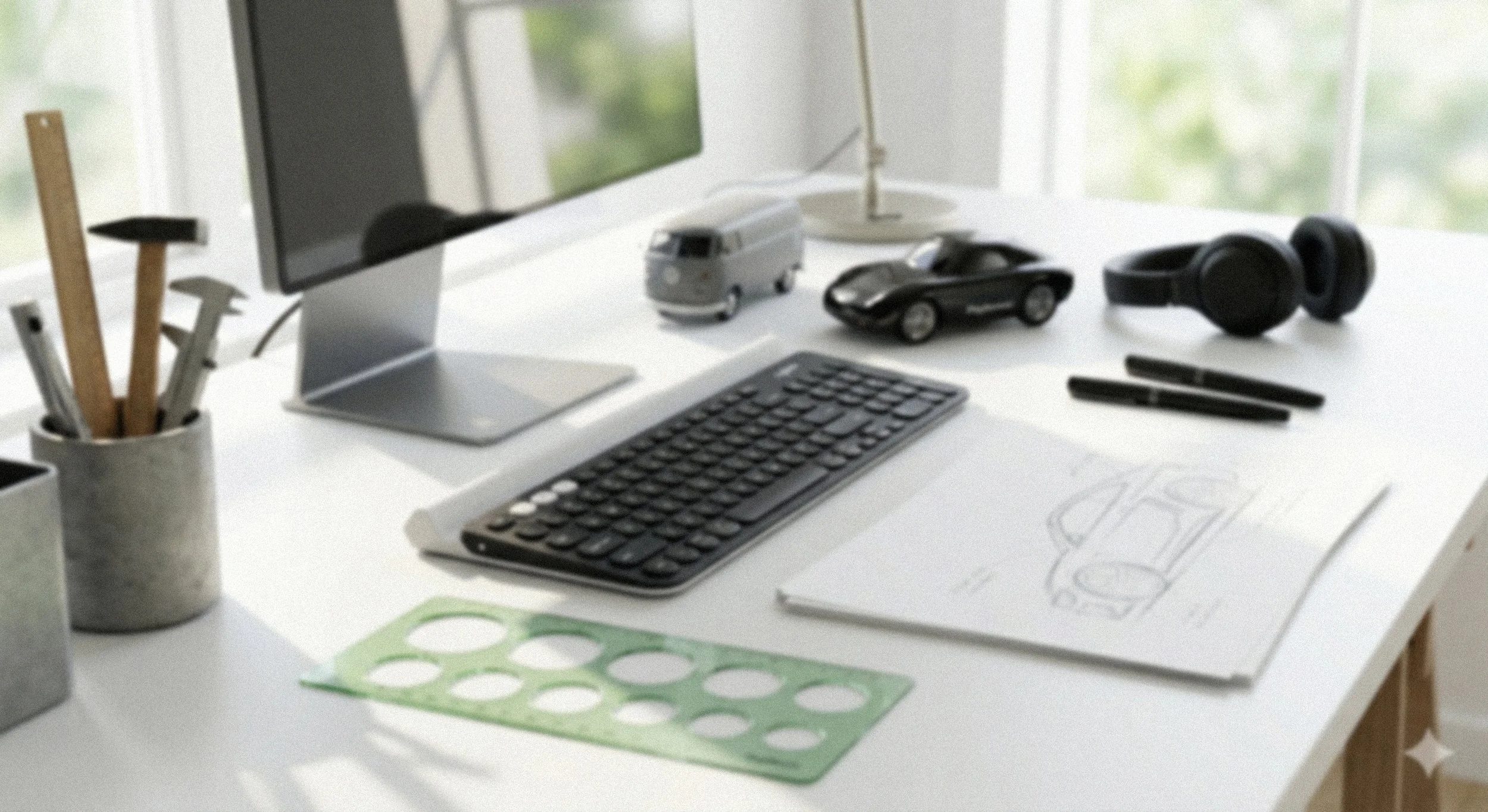 Clean white desk with computer monitor, keyboard, black pens, gray pencils in a container, small model camper van, black toy sports car, black headphones, sketch of a car, green stencil for circles, and a white desk lamp, with a window showing green trees in the background.