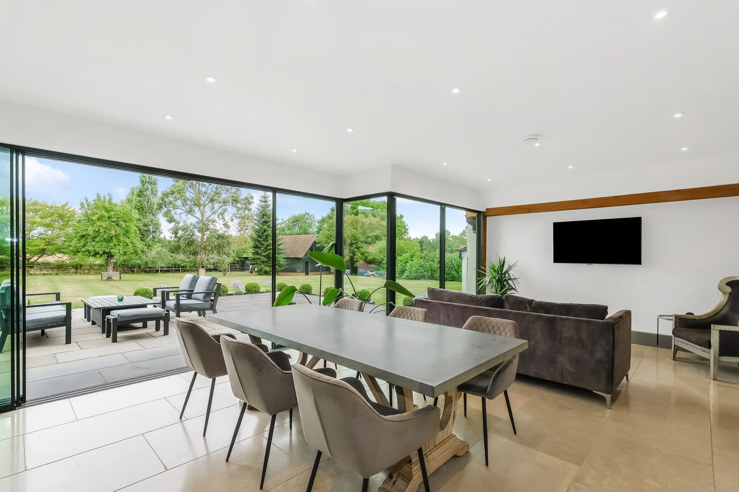 Modern living room with large glass sliding doors, outdoor patio with seating, green trees outside, white walls, and a mounted flat-screen TV.
