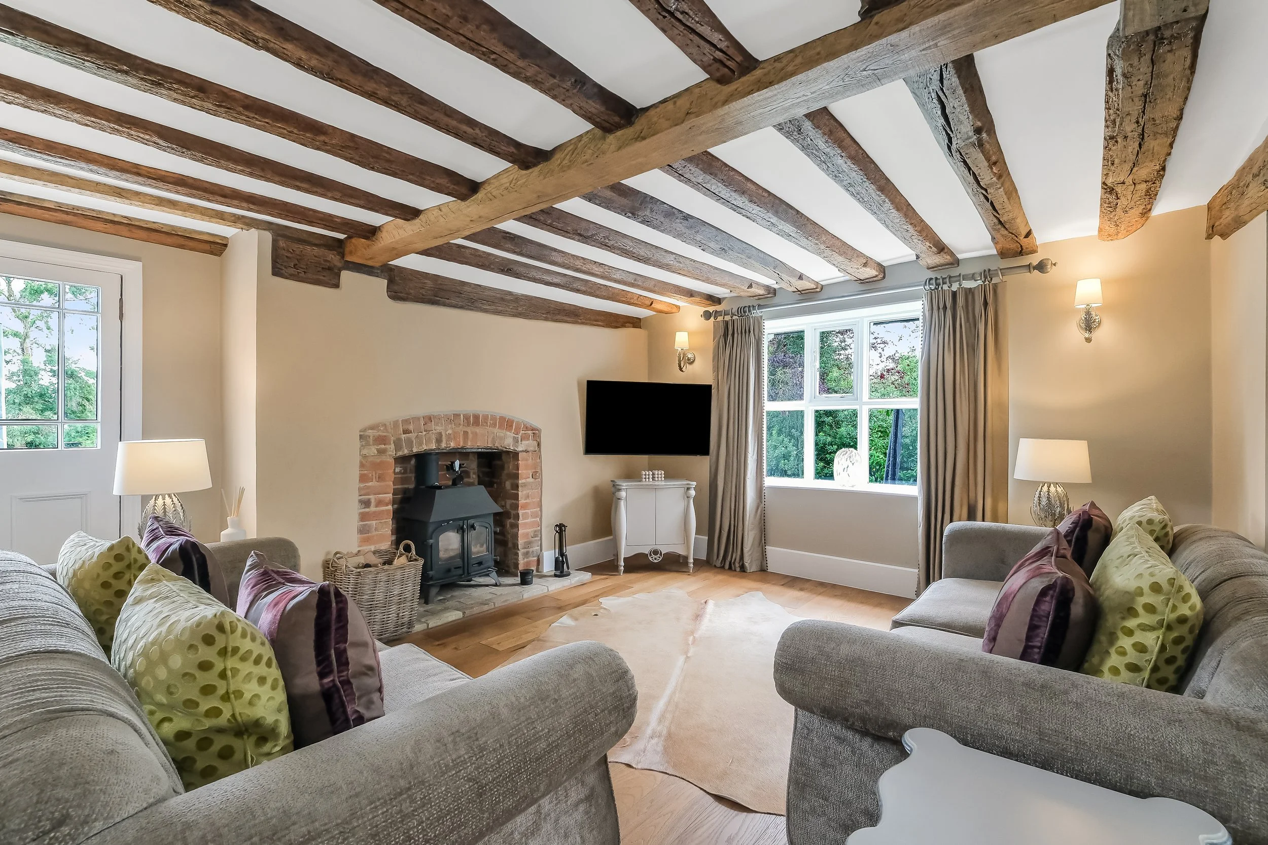 Living room with exposed wooden ceiling beams, beige walls, a fireplace, a wall-mounted TV, and chairs with pillows.