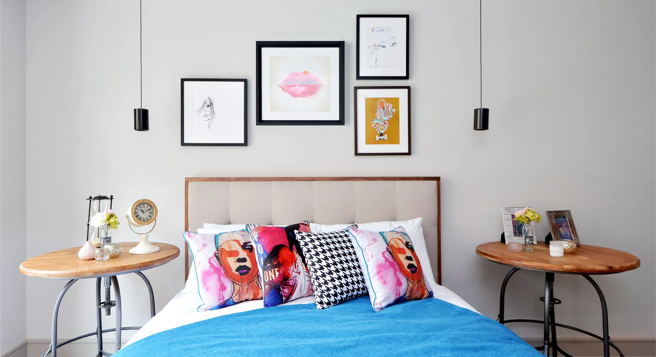 A bedroom with a bed, two wooden side tables, and a white wall decorated with five framed art pieces. The bed has colorful pillows, including one with a woman's face, and a blue blanket. The side tables hold decorative items like a clock, flowers, ca