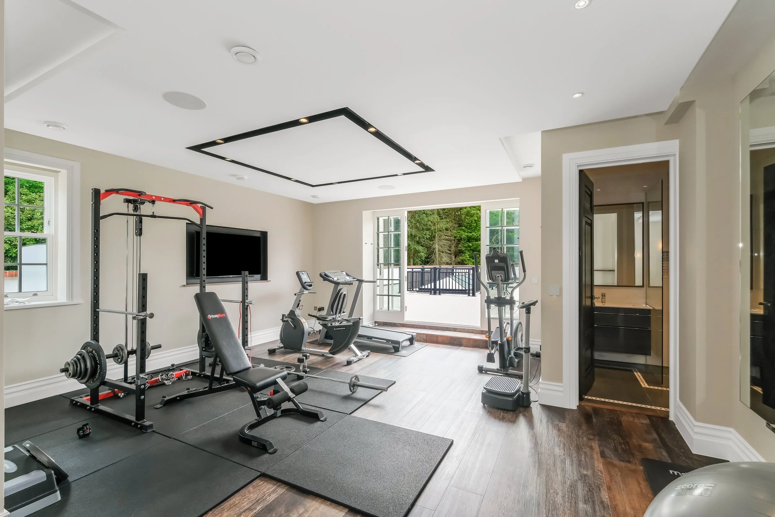 Home gym with exercise equipment including a treadmill, stationary bike, weightlifting bench, and a television, with large windows showing greenery outside.
