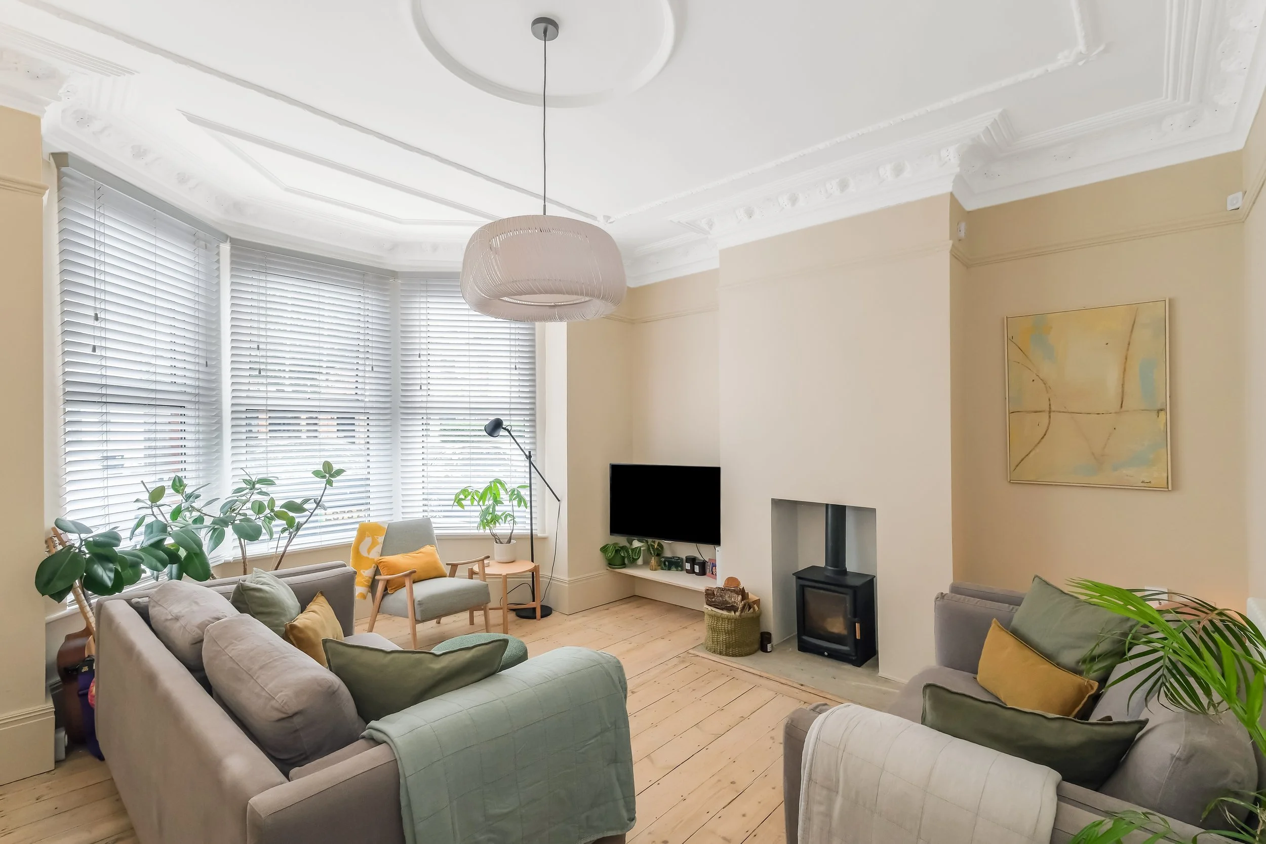 Living room with large window blinds, a television, a wood stove, and various seating including a green armchair, a gray sofa, and a gray armchair with yellow pillows. There are houseplants, a floor lamp, and artwork on the wall.