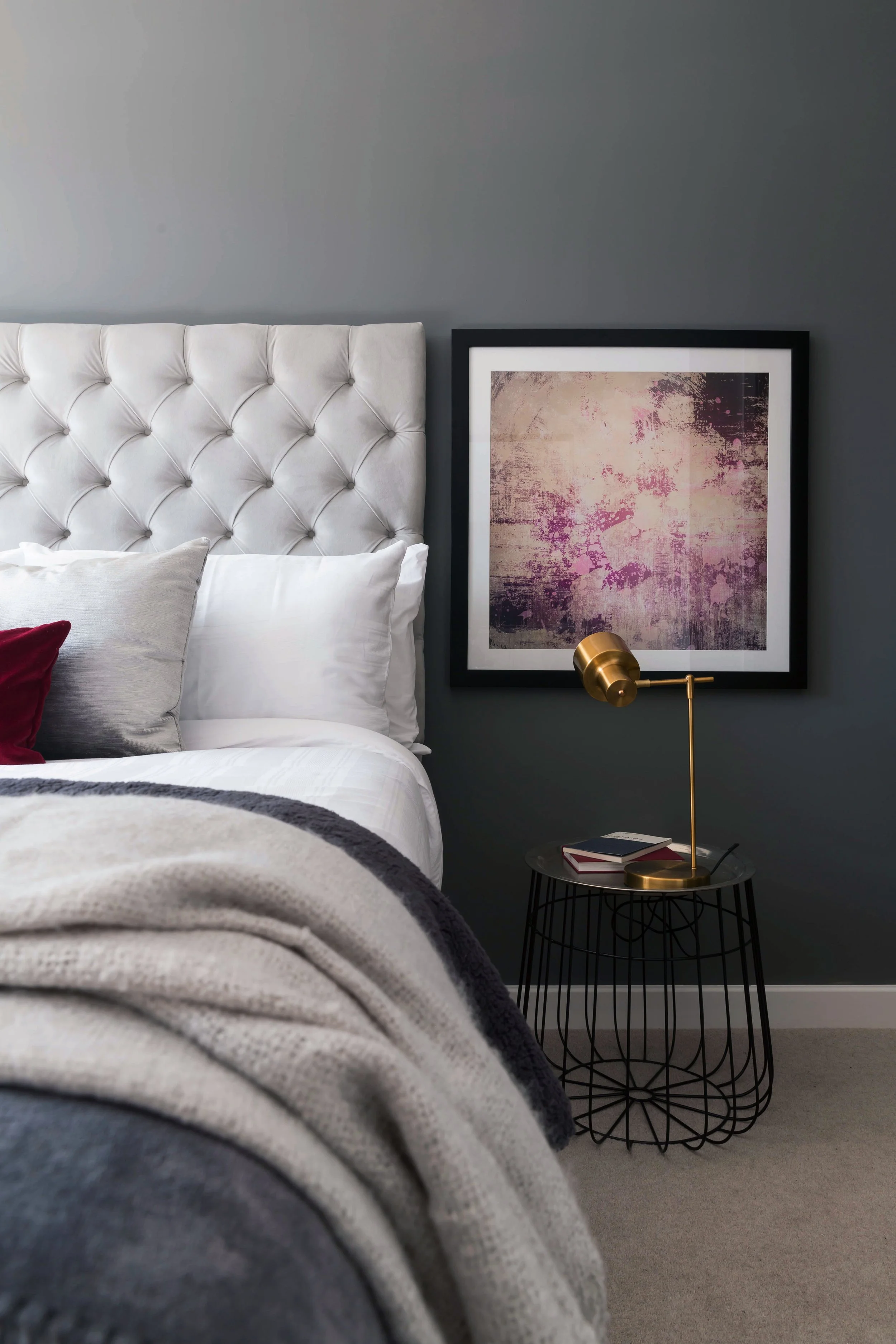 A bedroom with a white tufted headboard, white bedding, a gray blanket, and decorative pillows. A framed abstract art piece with pink, purple, and beige tones hangs on a dark gray wall. A black side table holds a gold desk lamp with a black base and 
