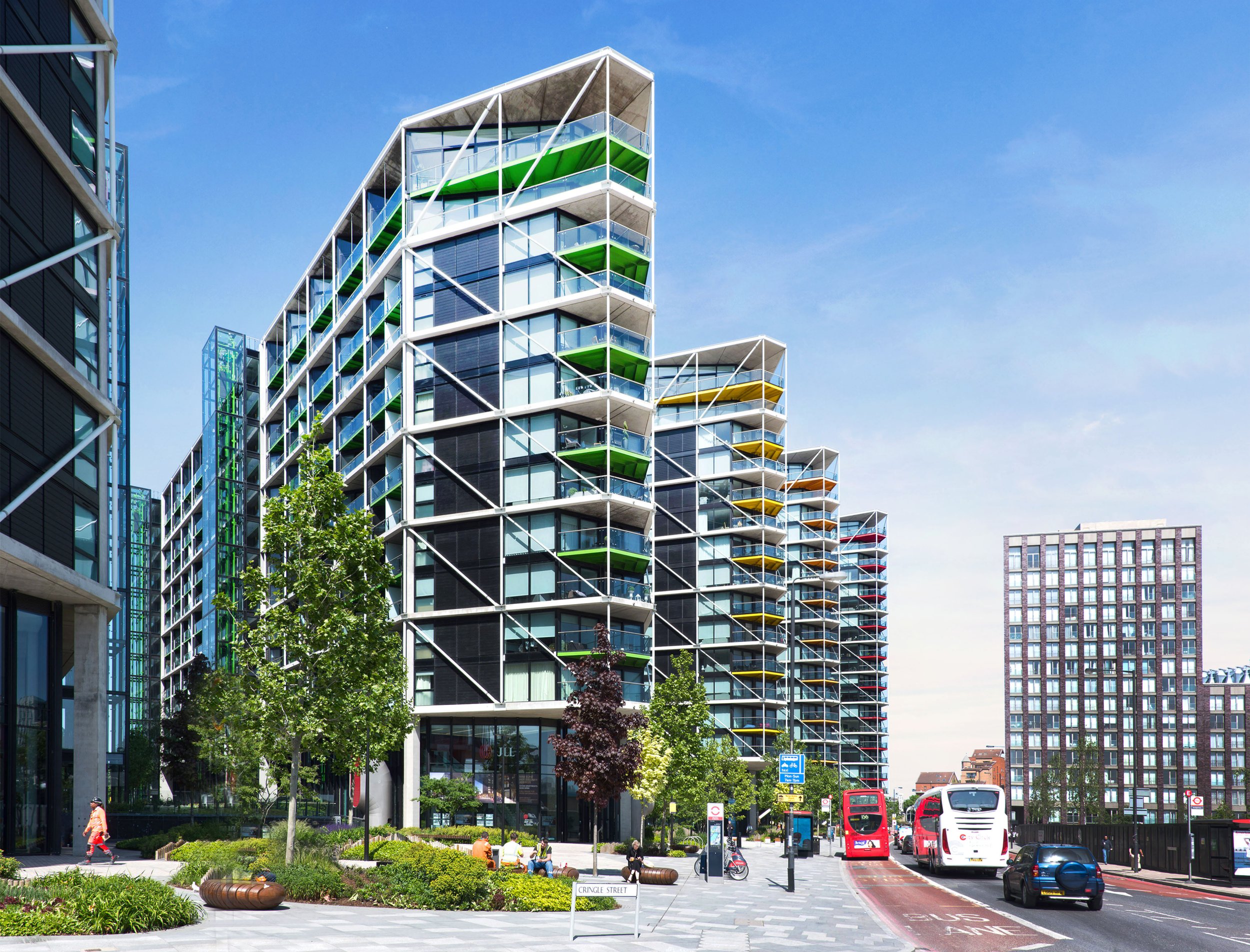 Modern residential building with colorful balconies on a sunny day, surrounded by trees and urban street scene with buses, cars, and people.