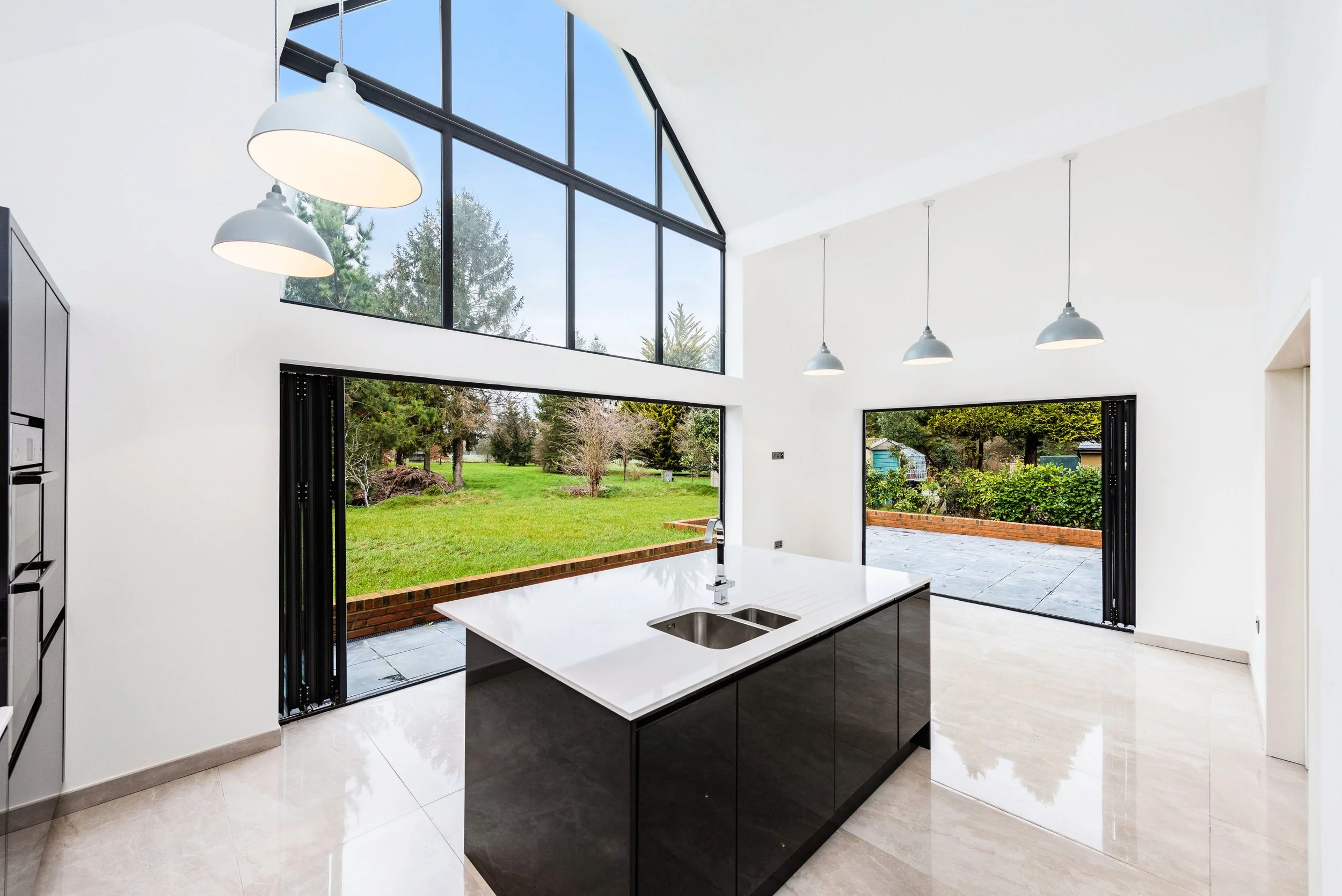 Modern kitchen with large glass windows and an outdoor view of a grassy yard and trees, featuring pendant lights and a white island with a sink.