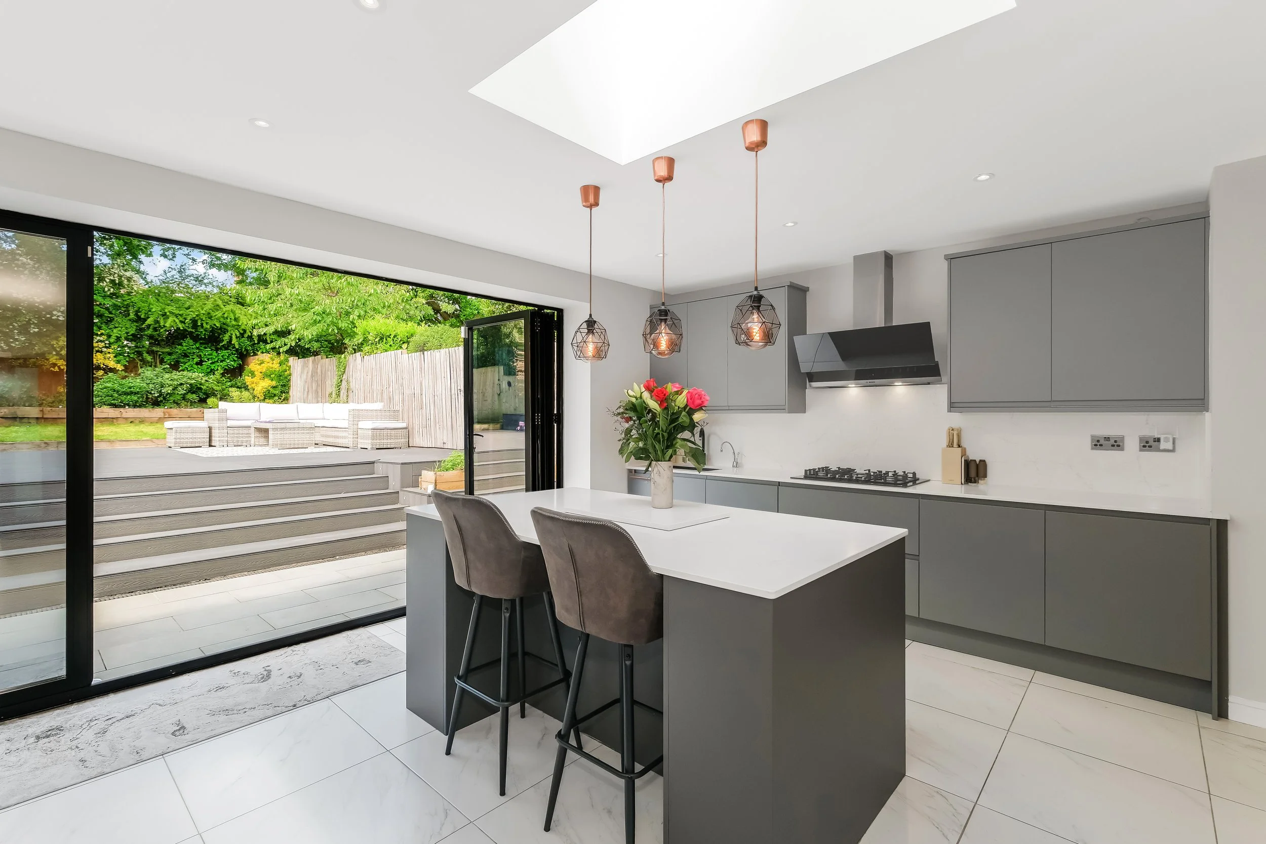 Modern kitchen with gray cabinets, white countertops, pendant lights, a vase of pink and white flowers, and an open view to a backyard patio with outdoor seating and greenery.