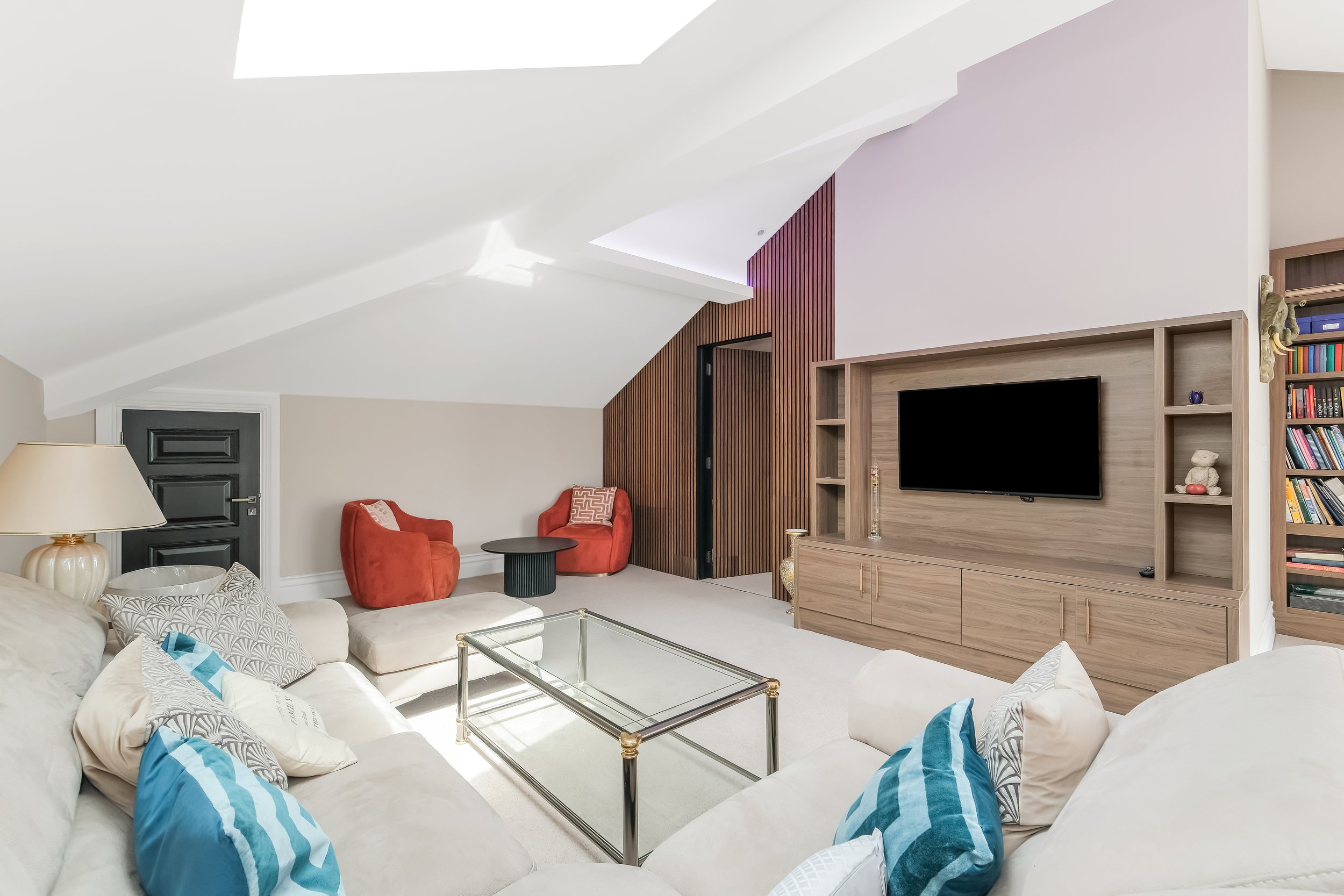 Living room with white sofa and blue striped pillows, two red armchairs, a black door, a wooden media console with a wall-mounted TV, built-in bookshelves, and decorative items on the shelves, with a large window letting in natural light.