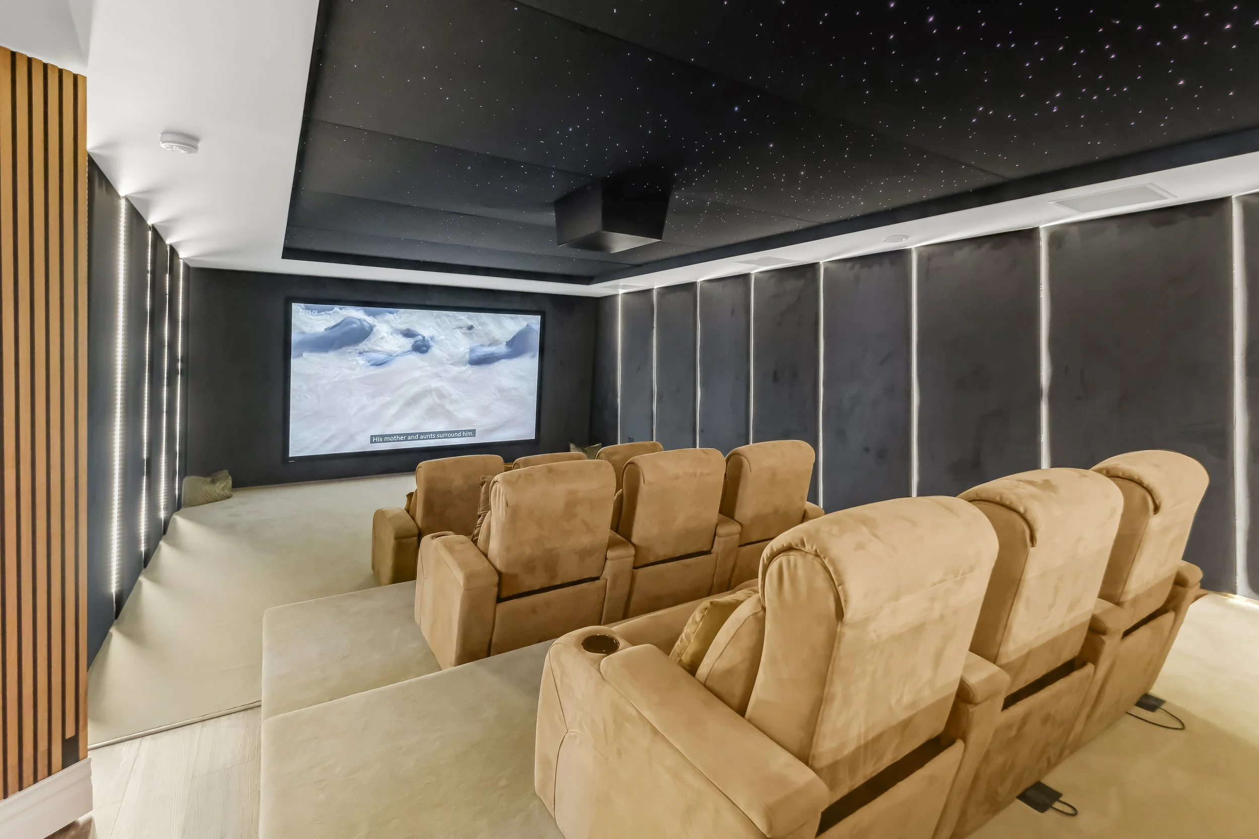 A cozy home theater room with multiple brown plush recliners facing a large flat-screen TV on a dark gray wall, with a ceiling decorated with star-like points.