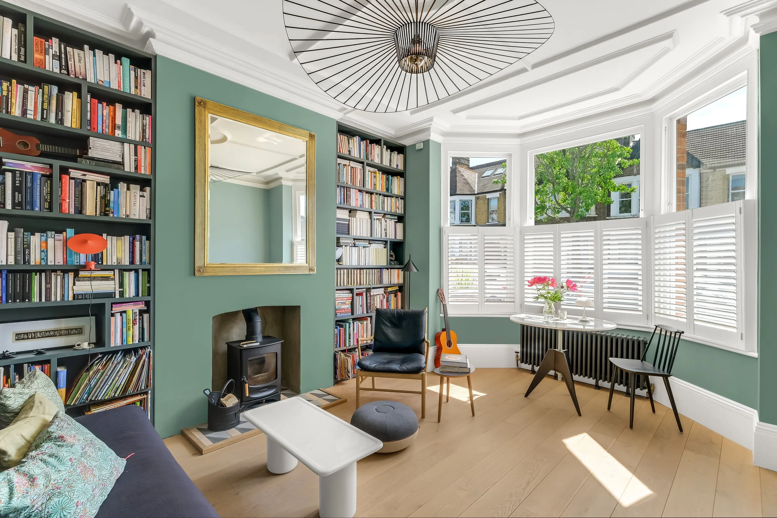 Bright living room with a large bay window with white shutters, green walls, a gold-framed mirror, bookshelves filled with books, a small wood stove, a guitar, a black armchair, a black dining chair, a small white coffee table, a round table with a p