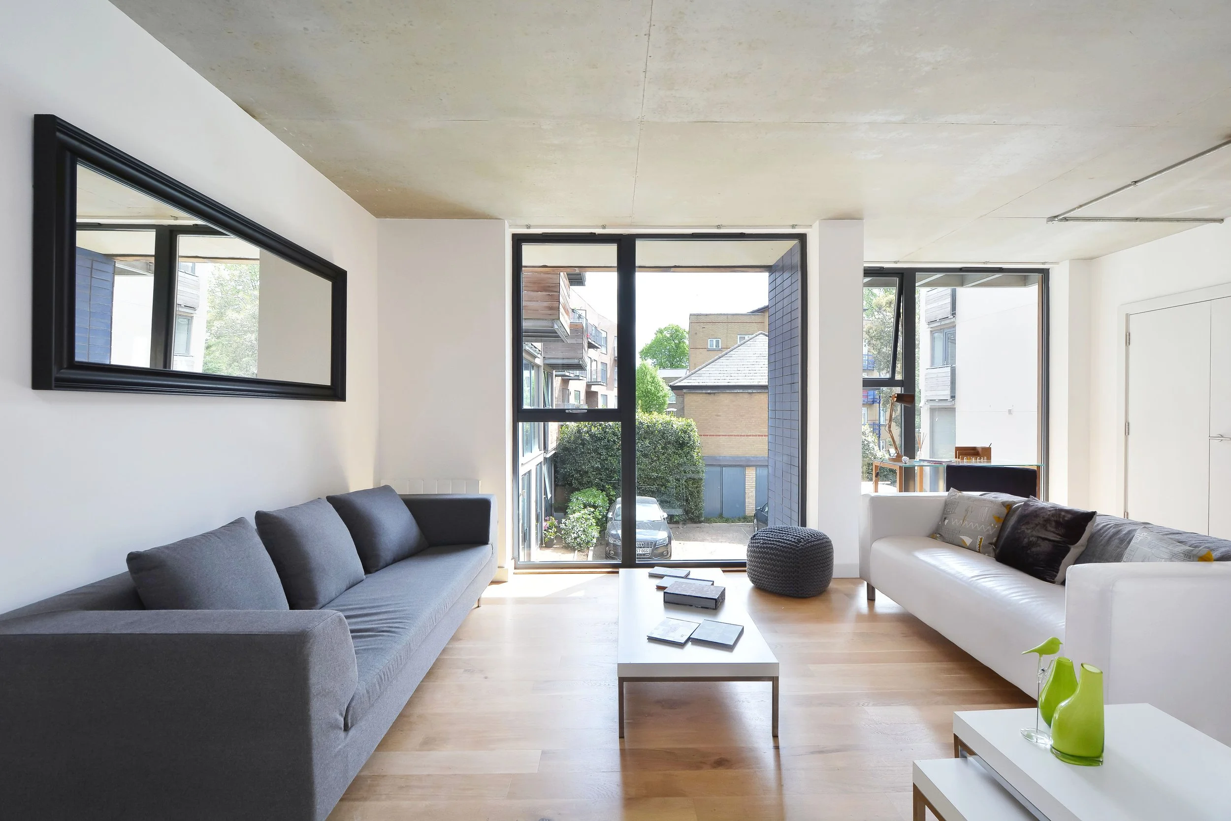A modern living room with a gray sofa on the left and a white sofa on the right, a small white coffee table in the center with books on it, large windows letting in natural light, and view of an outdoor parking area and other buildings.