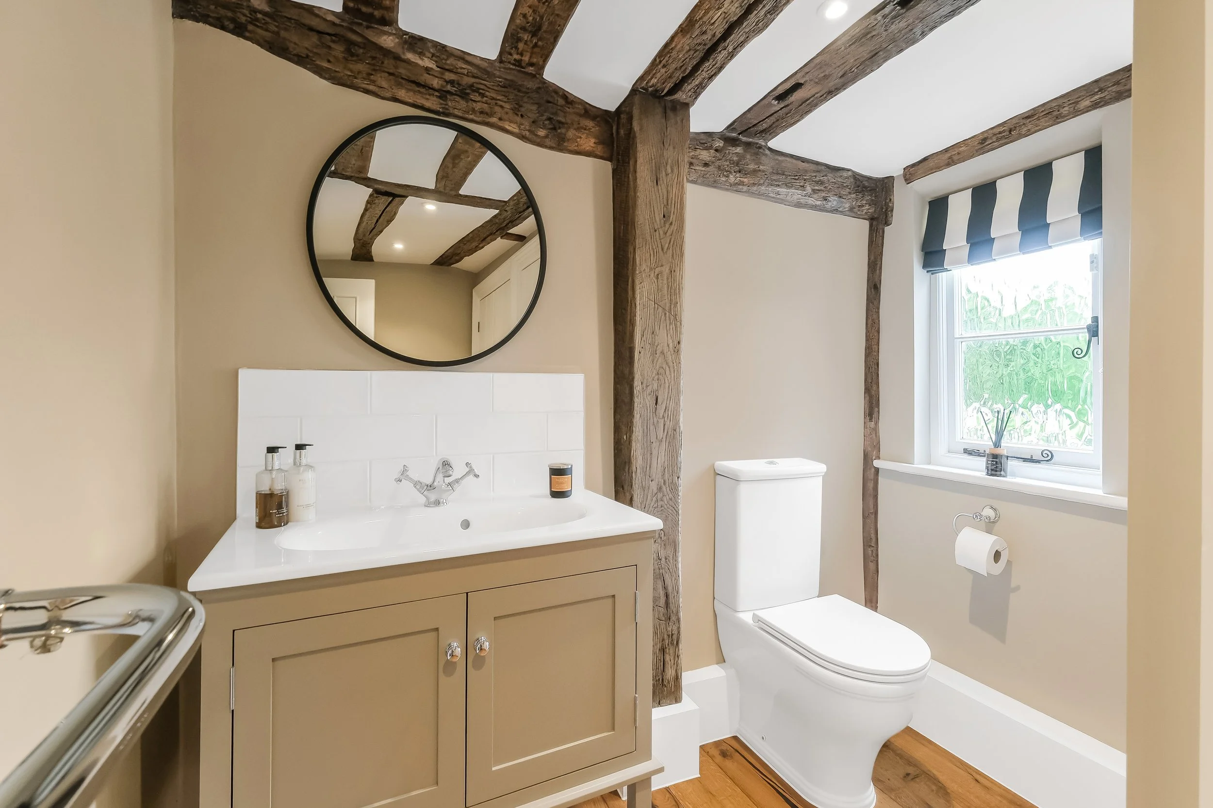 A bathroom with rustic wooden beams on the ceiling and walls, a beige vanity with a white sink, a round mirror, a white toilet, a window with a blue and gray striped Roman shade, and hardwood flooring.