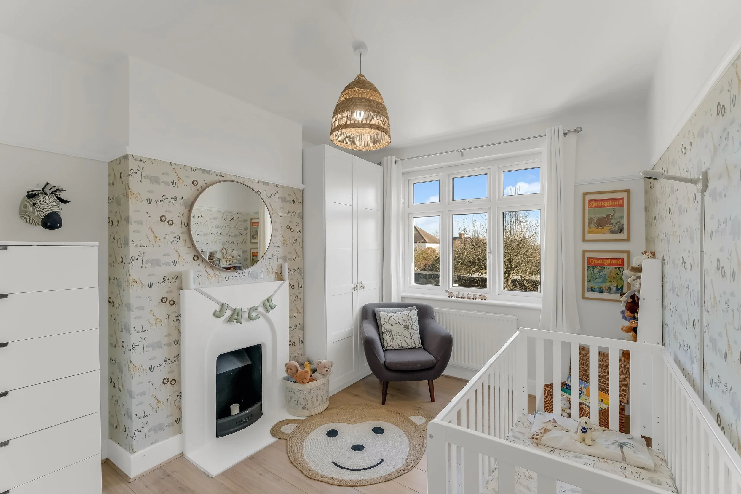 A nursery room with white walls decorated with animal themed wallpaper, a white closet, a grey armchair with a pillow, and a white crib with stuffed animals. There is a round mirror above a fireplace with a name banner 'JACK', and a small rug with a 