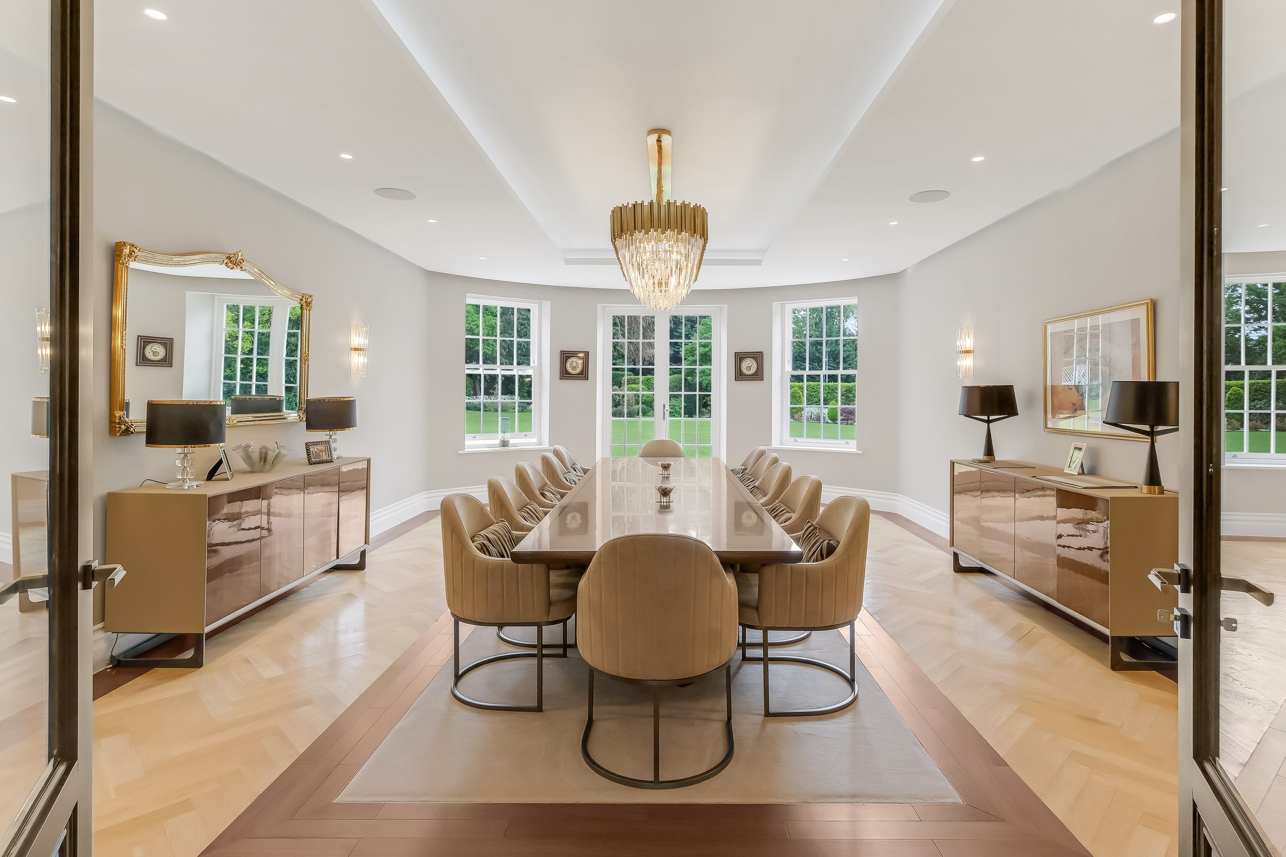 Elegant dining room with a large oval table, beige upholstered chairs, a large crystal chandelier, gold-framed mirrors, and windows overlooking greenery.