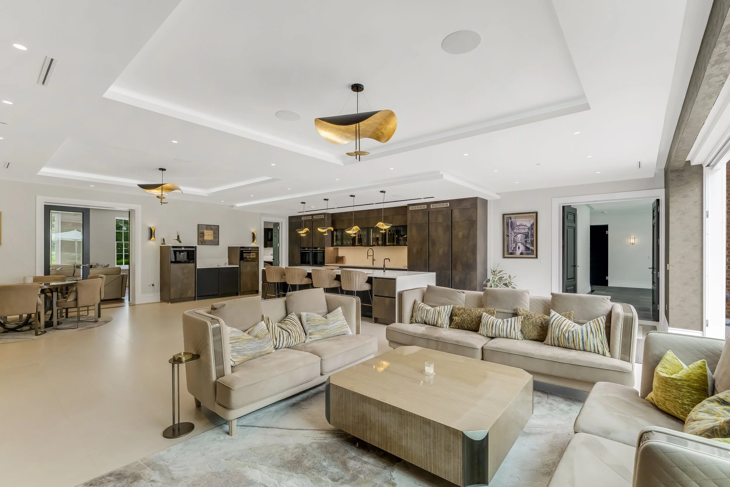 Spacious, modern living room with beige sofas, a large wooden coffee table, and a cozy area rug. The open kitchen features a white island and dark cabinetry with gold pendant lighting. Ceiling has recessed and decorative lighting, with large windows 